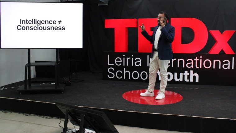 Fabiano Cruz stands on a red circular TEDx stage during a presentation at TEDx Leiria International School Youth. He wears a dark blazer, white shirt, light trousers, and white sneakers, holding a microphone and gesturing with one hand. Behind him are large red “TEDx” letters against a black backdrop. To the left, a screen displays the slide text: “Intelligence ≠ Consciousness.”